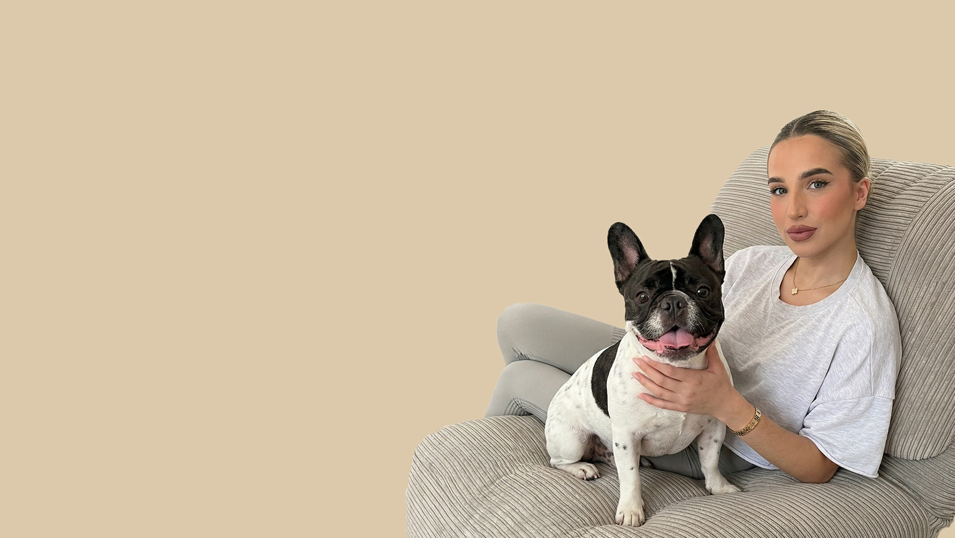 Woman sitting on a couch with a small black and white french bull dog, both looking at the camera against a beige background.
